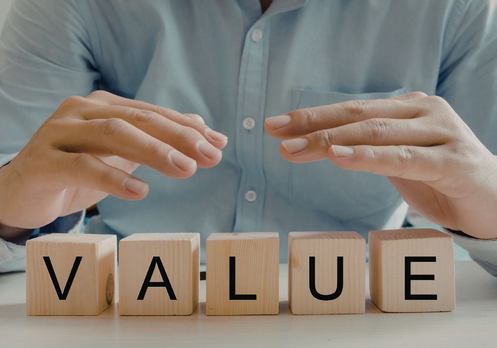 A person in a light blue shirt hovering their hands protectively over five wooden blocks that spell the word "VALUE" on a light-coloured surface.