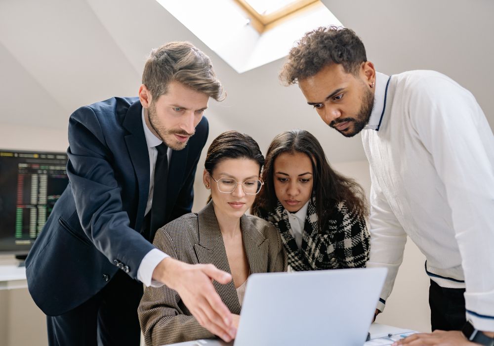 A group of four diverse young professionals gathered around a laptop in a modern office