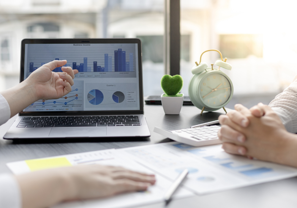 A close-up of a business meeting where one person points to a laptop screen displaying income bar charts and pie graphs