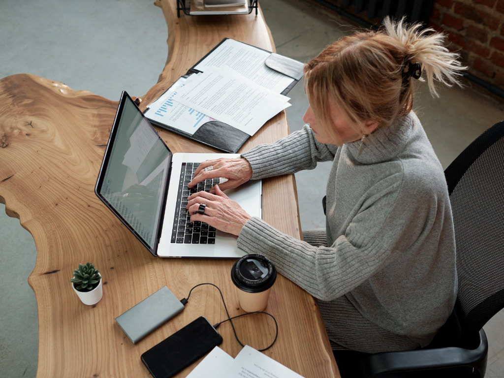 An elevated view of a woman with blonde hair in a grey turtleneck sweater typing on a laptop