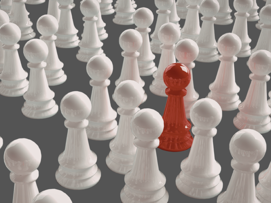 A single red chess pawn stands out prominently among a large crowd of uniform white pawns on a dark grey surface, symbolising individuality or leadership.