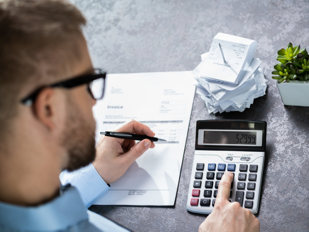 Over-the-shoulder view of a man with glasses using a calculator and holding a pen to review an invoice