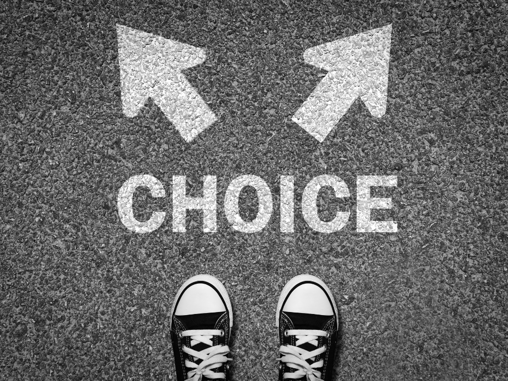 A top-down, black-and-white photo of a person's feet in sneakers standing on asphalt in front of the word "CHOICE" and two arrows pointing in opposite directions.