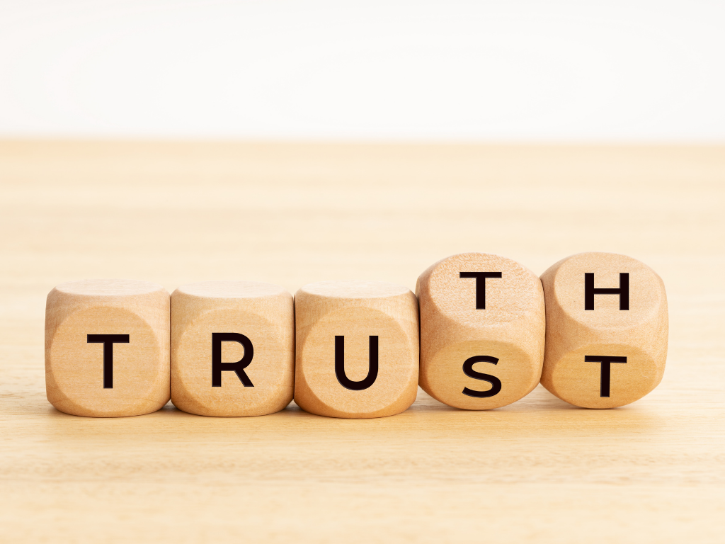 Five wooden blocks on a light surface arranged to spell "TRUST," with the final two blocks being rotated to also suggest the word "TRUTH."