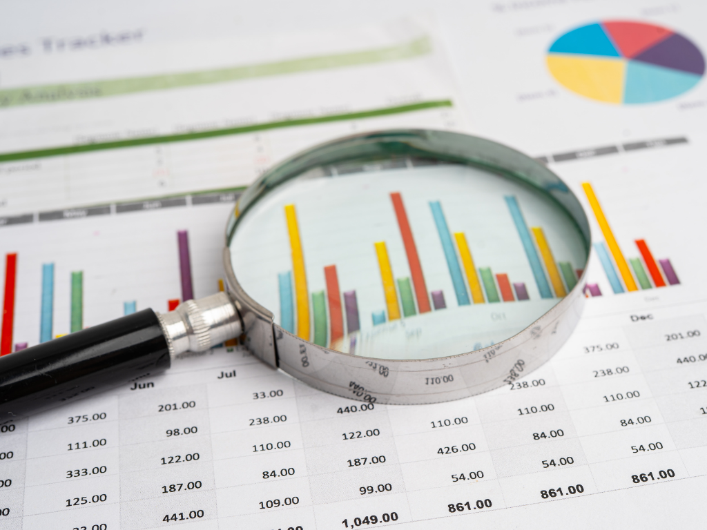 A magnifying glass rests on top of a financial report, enlarging a colourful bar chart and numerical data, with a pie chart visible in the background.