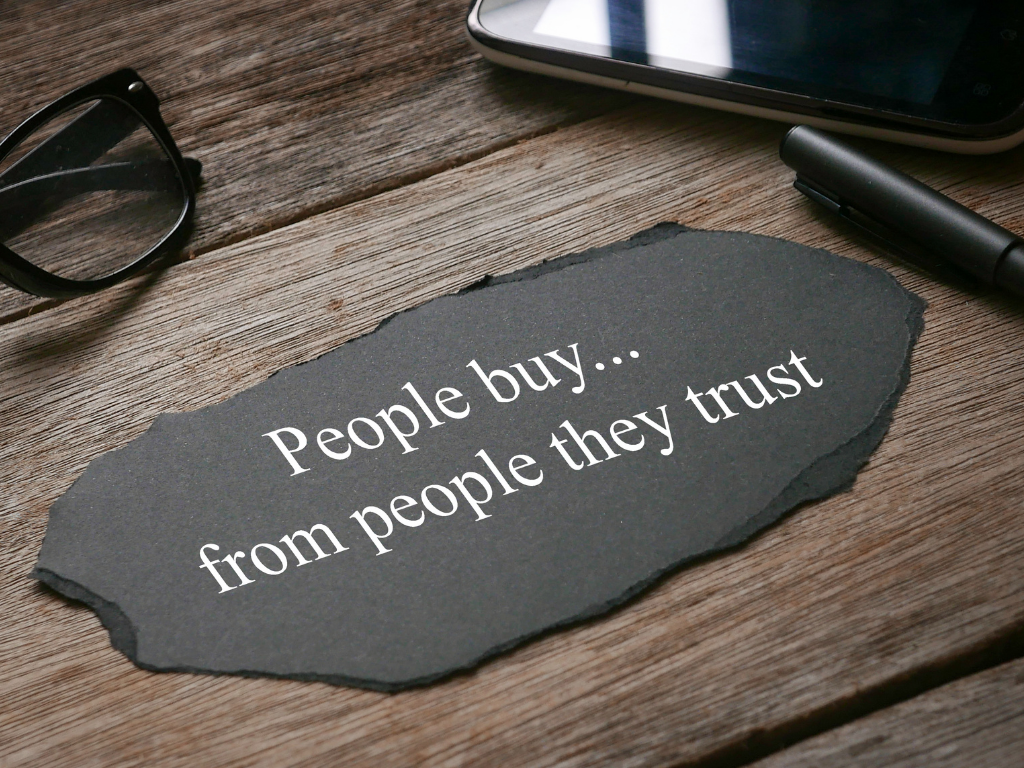 A piece of torn black paper with the white text "People buy... from people they trust" resting on a rustic wooden desk