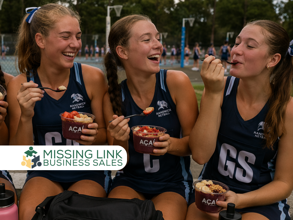 Three young women in Randwick Netball uniforms laughing while eating açaí bowls outdoors, with a "Missing Link Business Sales" logo in the bottom left corner.